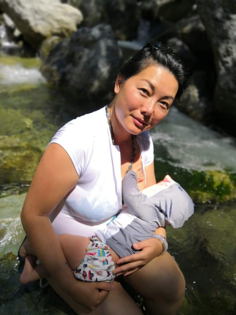Photo of a mother nursing her baby - she is sitting on a rock in a shallow river and looking towards the camera, smiling.